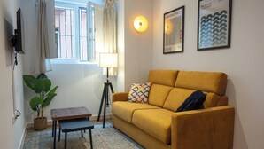A living room with a mustard-colored sofa, a small wooden table, a floor lamp, a potted plant, and two framed artworks on the wall.