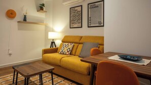 A cozy living room with a mustard yellow sofa, a wooden coffee table, and a small orange chair. There is a lamp, a wall-mounted shelf with a plant, and two framed artworks on the wall. - Apartments EL Aljibe - Ap.1 (Badajoz)