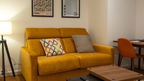 A yellow sofa with patterned cushions, a small wooden table, and a lamp. Two framed artworks are on the wall. - Apartments EL Aljibe - Ap.1 (Badajoz)