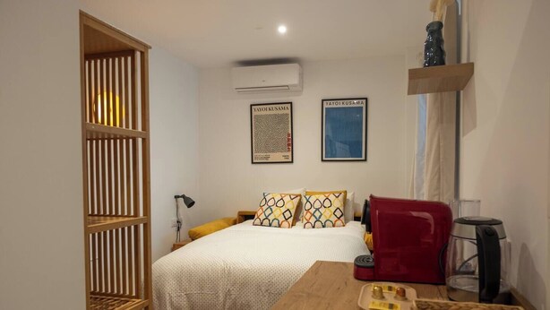 Un dormitorio con una cama, una mesita de noche de madera, una silla roja, una cafetera y dos cuadros en la pared.