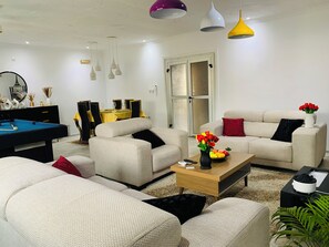 Ett modernt vardagsrum med ett biljardbord, beige soffor och en matplats med gula bordsdukar.