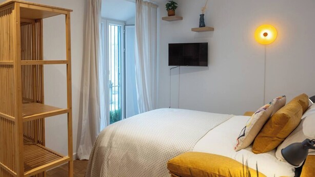 A bedroom with a bed, a wooden shelf unit, a TV, and a wall-mounted light. - EL Aljibe Apartments - Ap.4 (Badajoz)