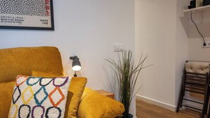 A living room with a mustard-colored sofa, a patterned throw pillow, a floor lamp, a tall plant in a black pot, and a wooden chair. - EL Aljibe Apartments - Ap.4 (Badajoz)