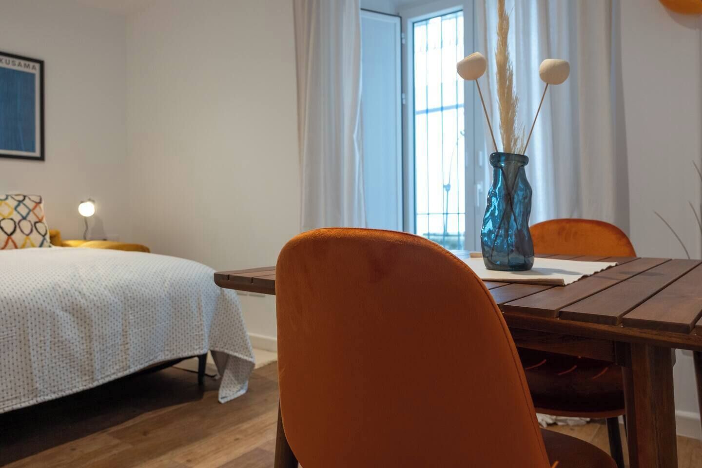 A hotel room with a bed, a dining table with chairs, a vase with dried plants, and a window with curtains.