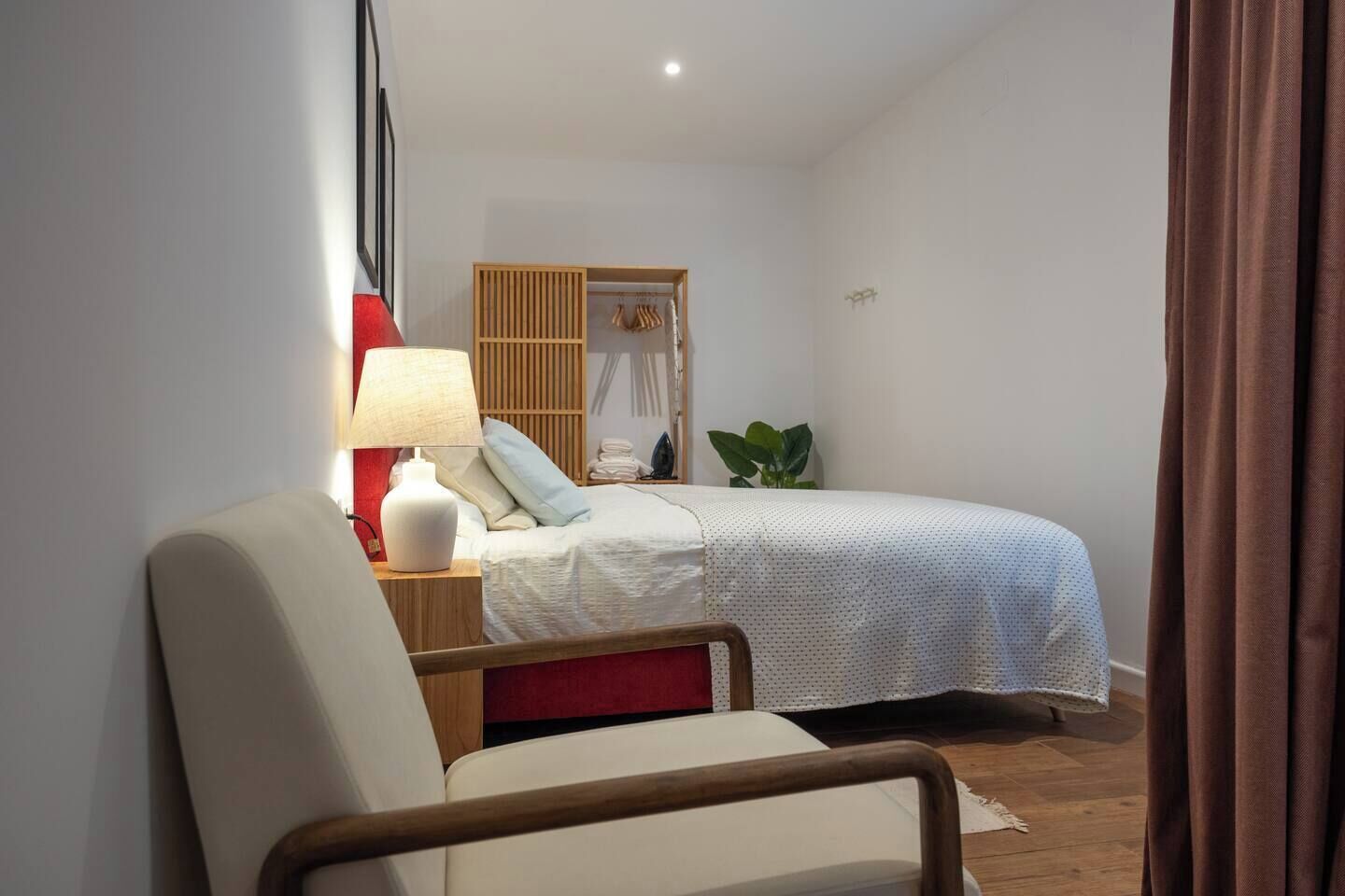 A bedroom with a bed, a chair, a nightstand with a lamp, a wooden wardrobe, and a potted plant.