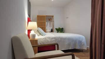 A bedroom with a bed, a chair, a nightstand with a lamp, a wooden wardrobe, and a potted plant.