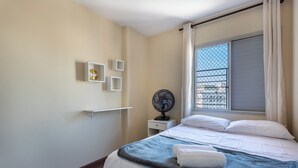 Un lit bien fait, avec du linge de lit blanc, une couverture bleue et une serviette pliée. On aperçoit une petite table de chevet blanche, sur laquelle se trouve un ventilateur, et un objet décoratif est accroché au mur.