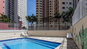 Une piscine entourée d’une clôture blanche, bordée d’un immeuble doté de balcons et orné d’une fresque murale.