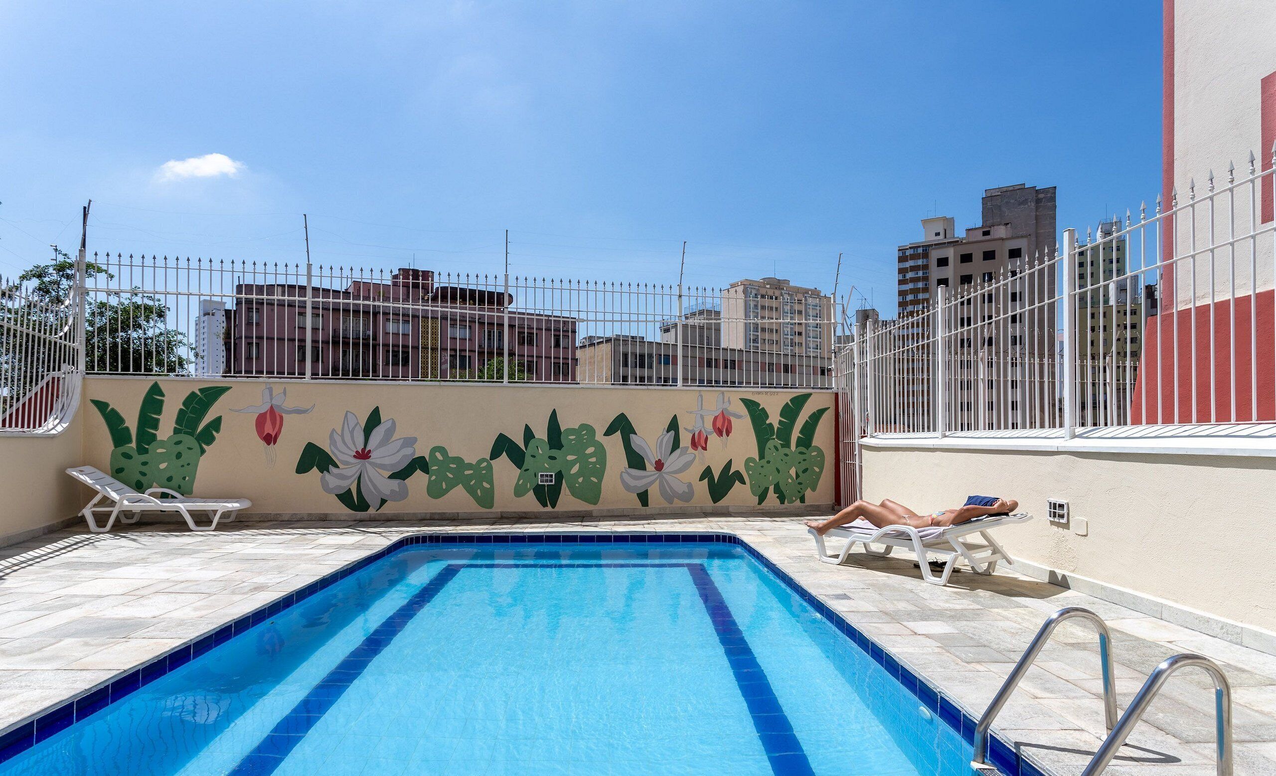 Une piscine sur le toit, bordée de carreaux, ornée d’une fresque représentant des plantes tropicales et entourée d’une clôture blanche.