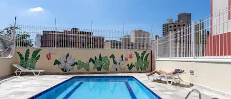 Una piscina en la azotea con borde de azulejos, un mural de plantas tropicales y una cerca blanca.