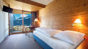 A hotel room with a bed, a TV, a table with chairs, and a view of mountains.