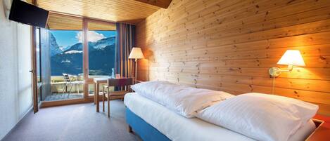 Habitación de hotel con cama, televisión, una mesa con sillas y vista a las montañas.