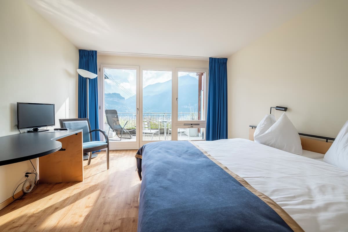 Habitación de hotel con cama, escritorio con televisor, silla y balcón con vistas a montañas.