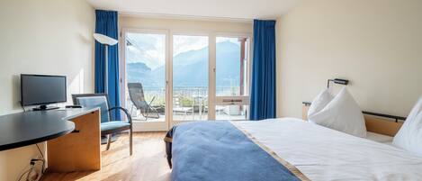 A hotel room with a bed, a desk with a TV, a chair, and a balcony with a view of mountains.