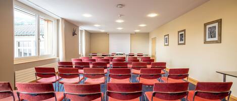 Une salle de conférence avec des rangées de chaises rouges, un écran de projection et une grande fenêtre équipée de stores.