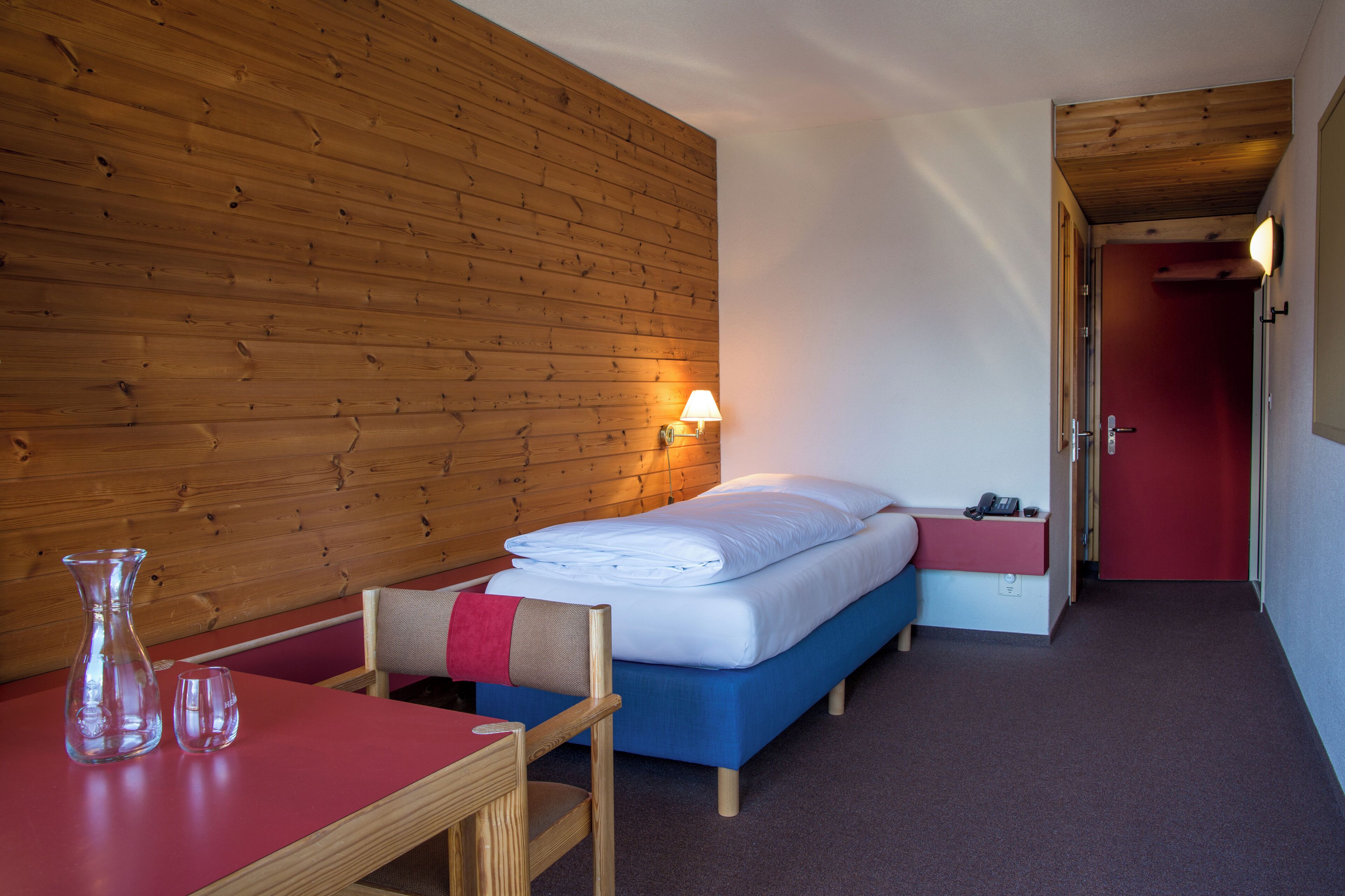 Ein Zimmer mit einer Holzwand, einem Bett mit blauer Matratze, einer roten Tür und einem kleinen Tisch mit einer Vase und Gläsern.