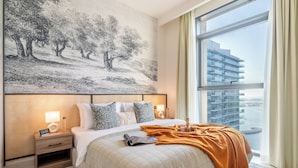 Une chambre d’hôtel moderne avec un grand lit, une table de chevet avec une lampe, un tableau représentant des arbres et une vue sur un immeuble par la fenêtre.