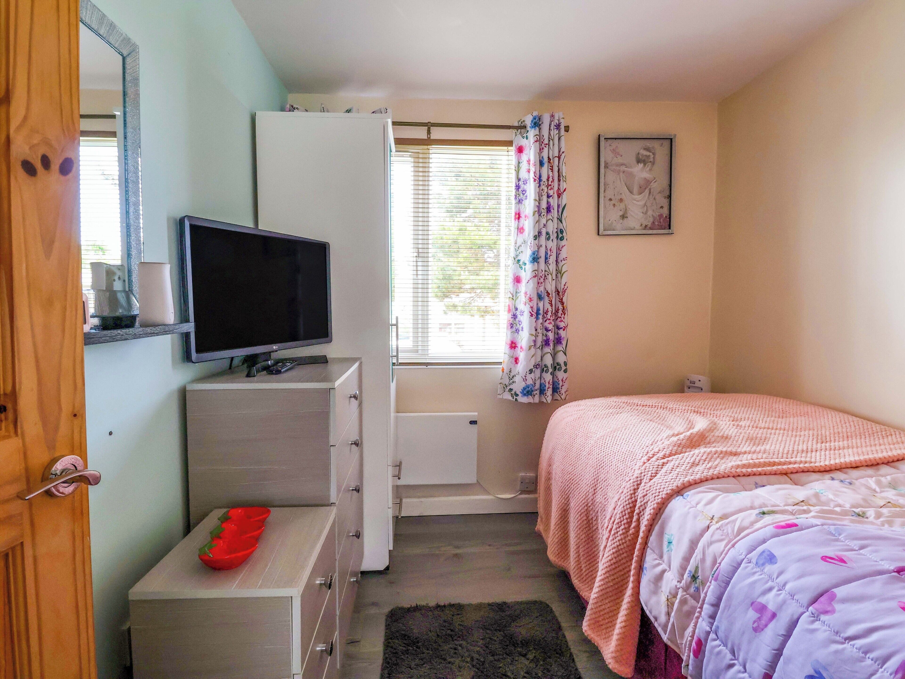 Ein Schlafzimmer mit Bett, Fernseher auf einer Kommode, einem Fenster mit Vorhängen und einer Heizung.