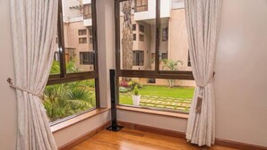 Una habitación con ventanas amplias, suelo de madera y vistas a un jardín y edificios exteriores.