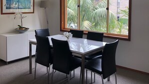 A dining area with a table and chairs, a white cabinet, a framed picture, and a window with a view of palm trees.