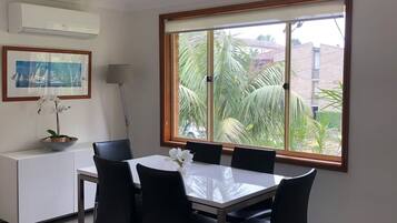 A dining area with a table and chairs, a white cabinet, a framed picture, and a window with a view of palm trees.