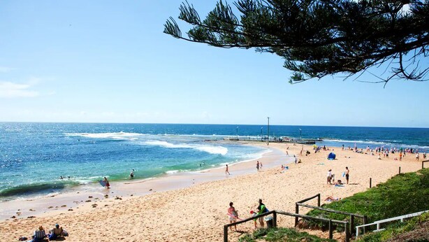 A beach with people sunbathing and swimming, a wooden walkway, and a clear blue sky.