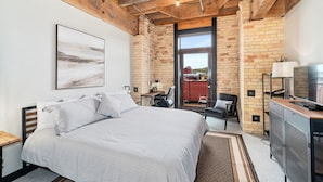A bedroom with a large bed, a desk, a chair, a TV, and a view of the outside.