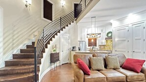 Ein geräumiges Wohnzimmer mit einer geschwungenen Treppe, einem Ledersofa mit roten Kissen und einer Küche im Hintergrund.