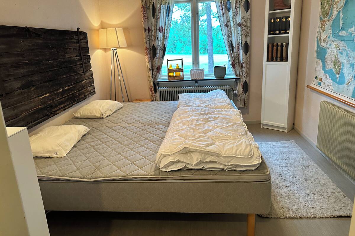 A bedroom with a bed, a lamp, a radiator, a window with curtains, a map on the wall, and a bookshelf.