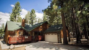 A log cabin with a porch, stone garage, and surrounded by tall pine trees. - Luxury w Spa Foosball - DeCarlo House (Big Bear Lake)