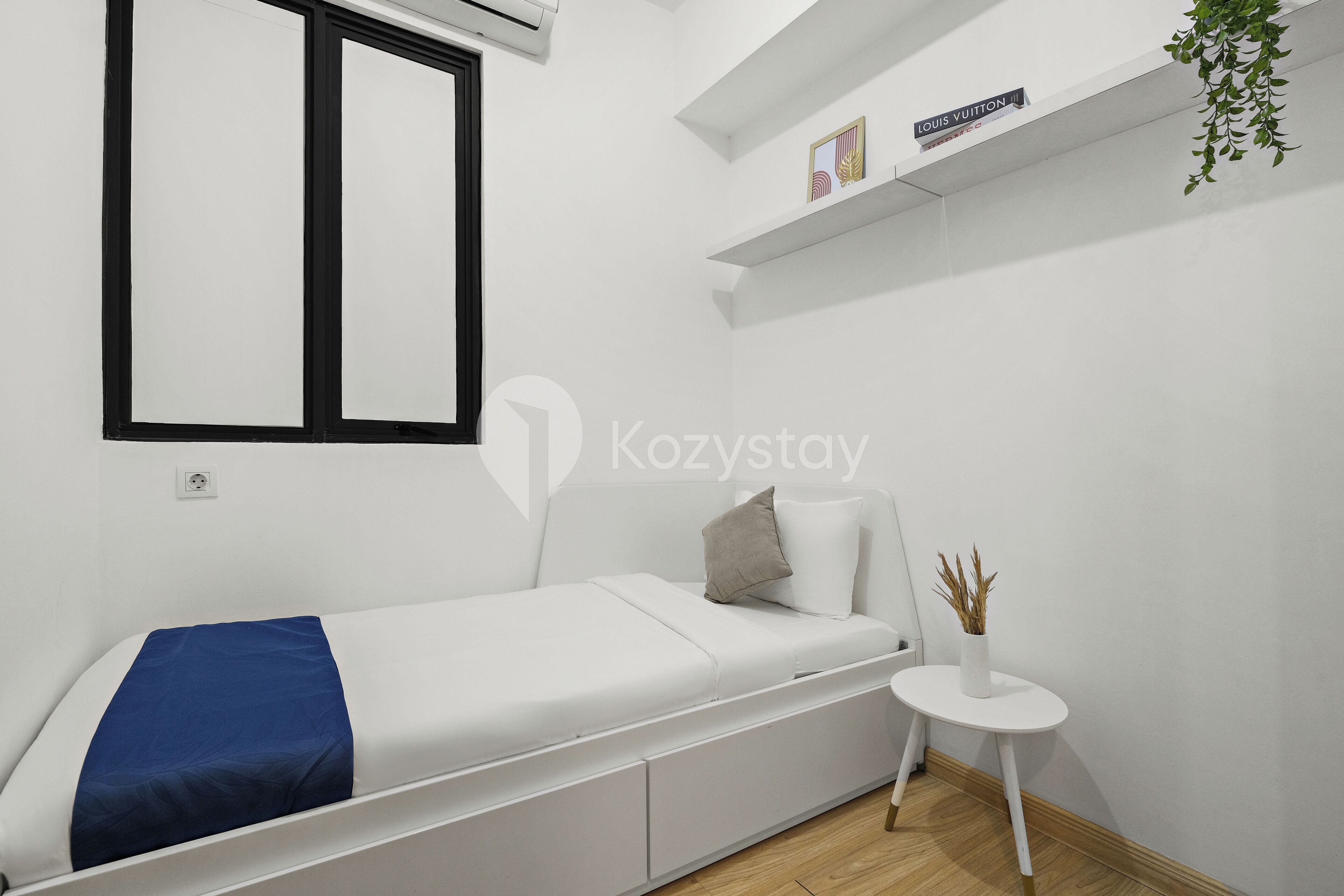 Une petite chambre moderne, avec un lit simple, une petite table, une fenêtre et une étagère ornée d’un logo Louis Vuitton.