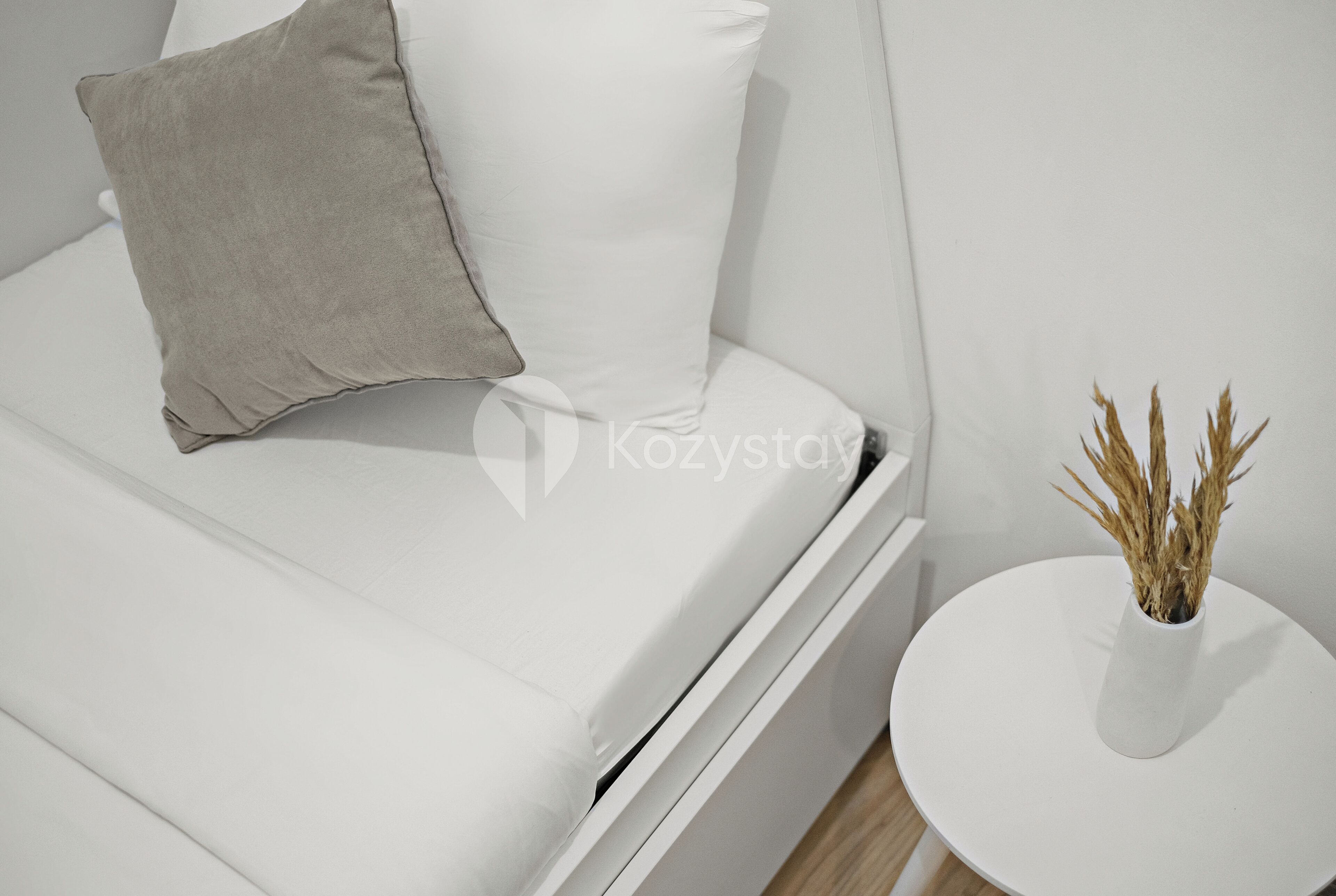 Un sofá blanco con una almohada gris y un jarrón blanco con plantas secas sobre una mesita auxiliar blanca.