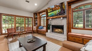 A spacious living room with a fireplace, large windows, and a television mounted above it. - Birdsong - Hot Tub, Lawn Games, Close to Wineries (Fredericksburg)