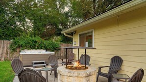 Un patio al aire libre con un pozo de fuego de piedra rodeado de sillas Adirondack, una mesa de madera y una parrilla de barbacoa empotrada.