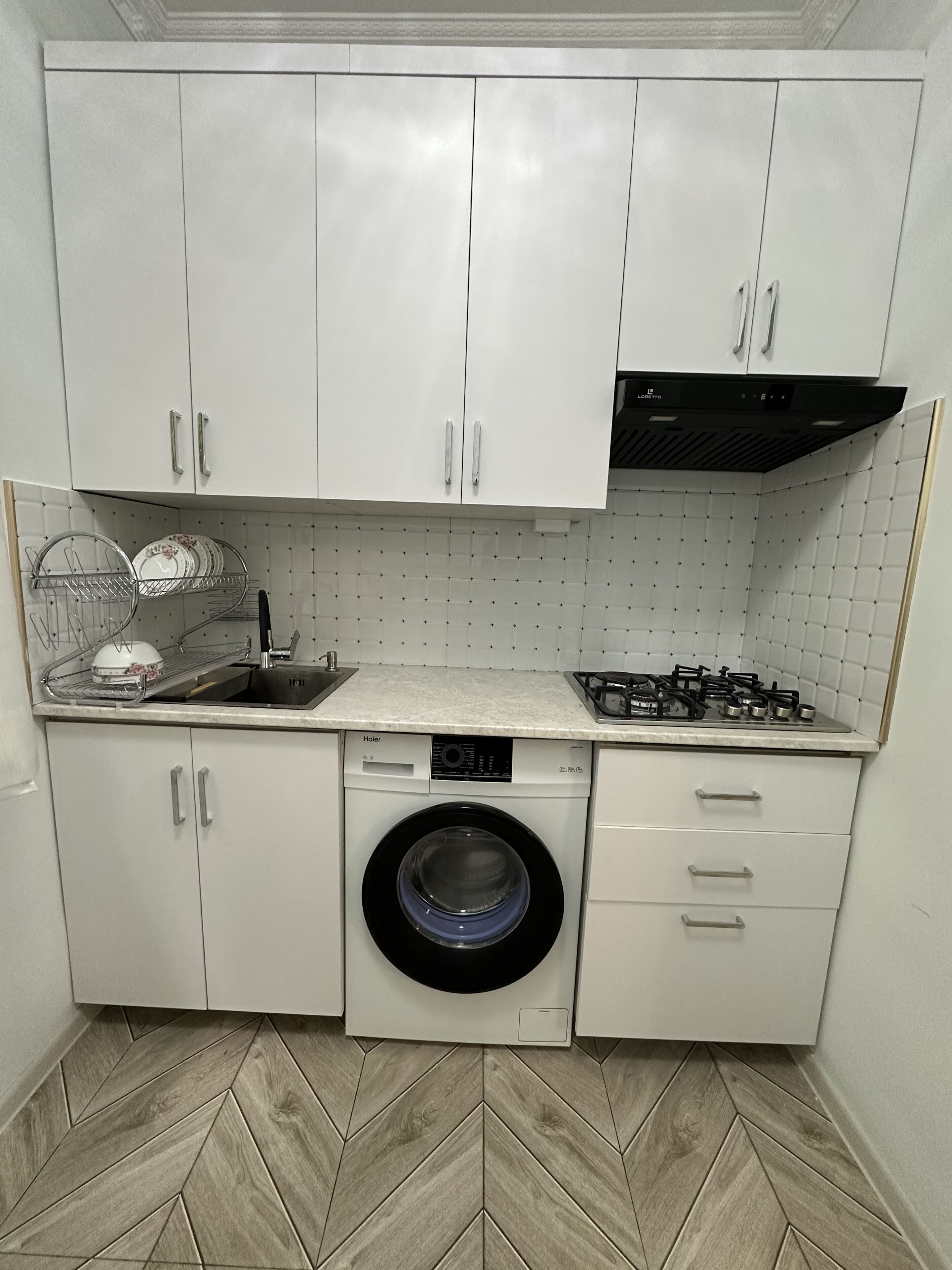 Cocina compacta con alacenas blancas, lavadora integrada, estufa a gas y salpicadero de azulejos.
