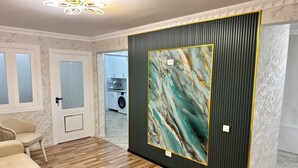 Huoneessa on puulattia, beigenvärinen sohva, lasipaneeleilla varustettu ovi ja suuri seinälle ripustettu taideteos.