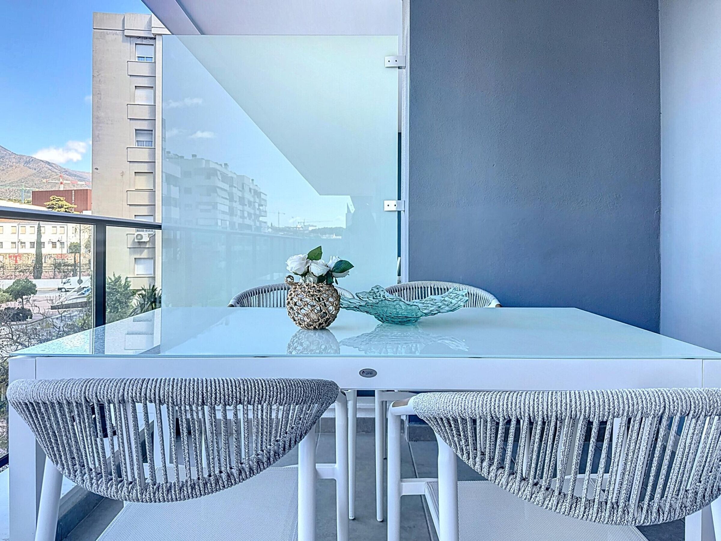 Een modern balkon met een glazen tafel, rieten stoelen en een decoratieve schaal.