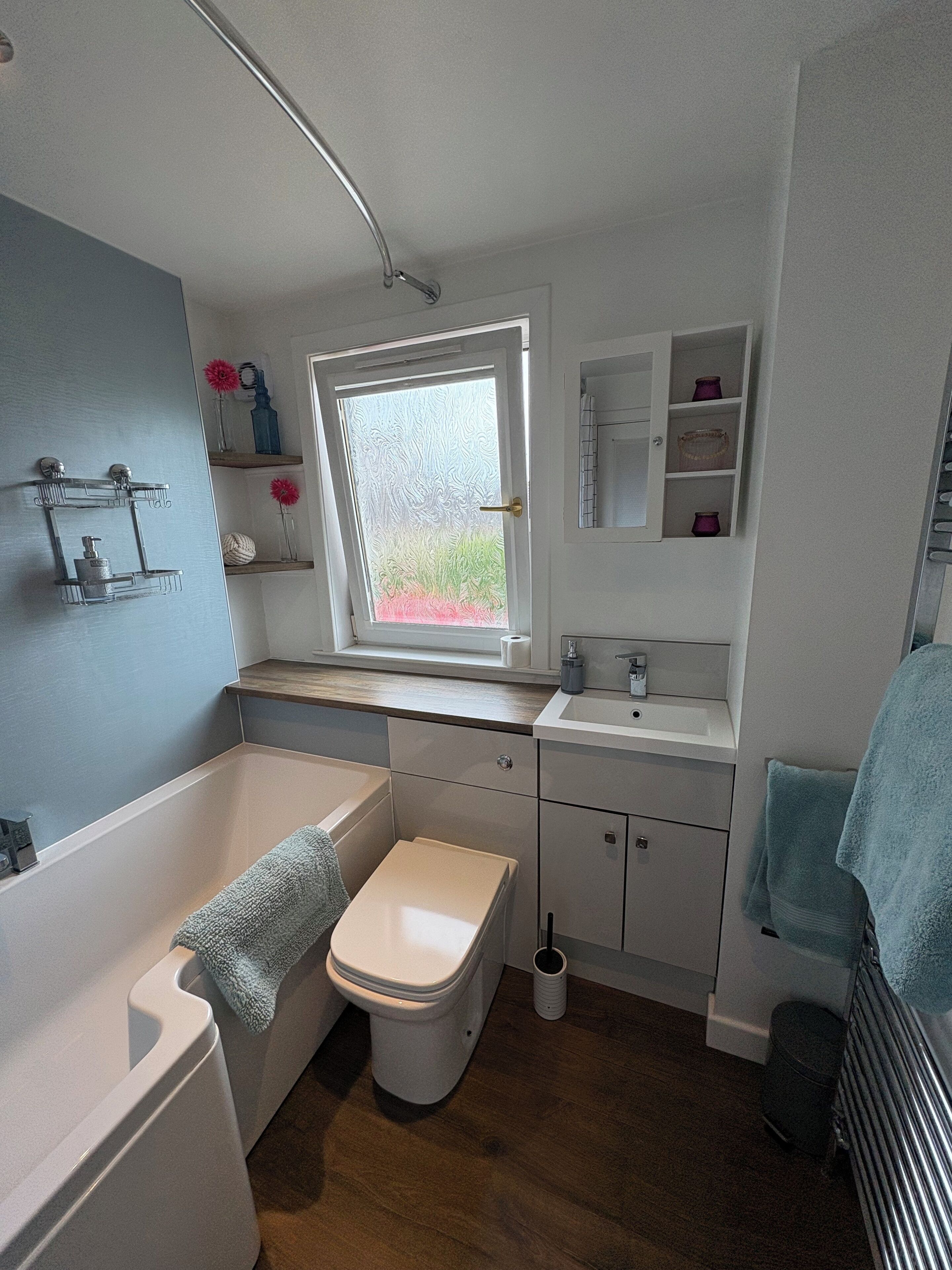 A compact bathroom with a bathtub, toilet, and sink. There is a window with frosted glass and a view of greenery outside.
