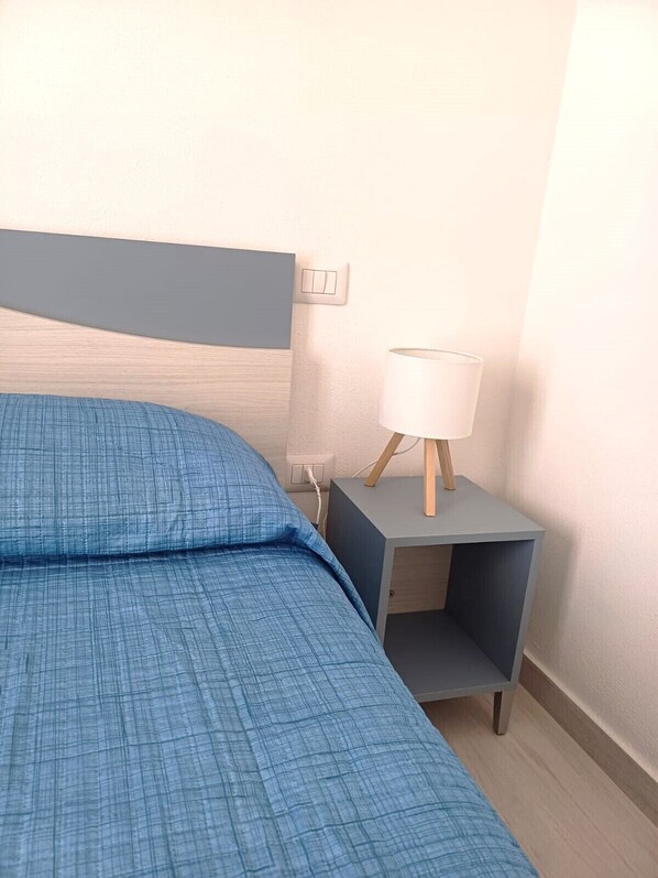 A neatly made bed with a blue checkered bedspread, a bedside table with a lamp, and a power outlet on the wall. - Affittimoderni La Maddalena - MADA43 (La Maddalena)