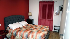 A bedroom with a tufted headboard, a plaid bedspread, a bedside table with a lamp, a small stool, and a red curtain. - Appia Antica Room (Marino)