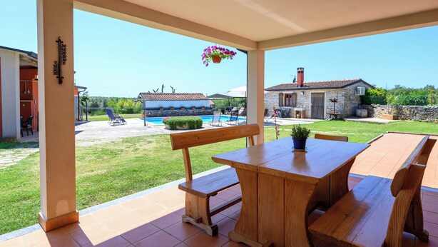 Une terrasse couverte avec des meubles en bois, une piscine et, au fond, des bâtiments en pierre.