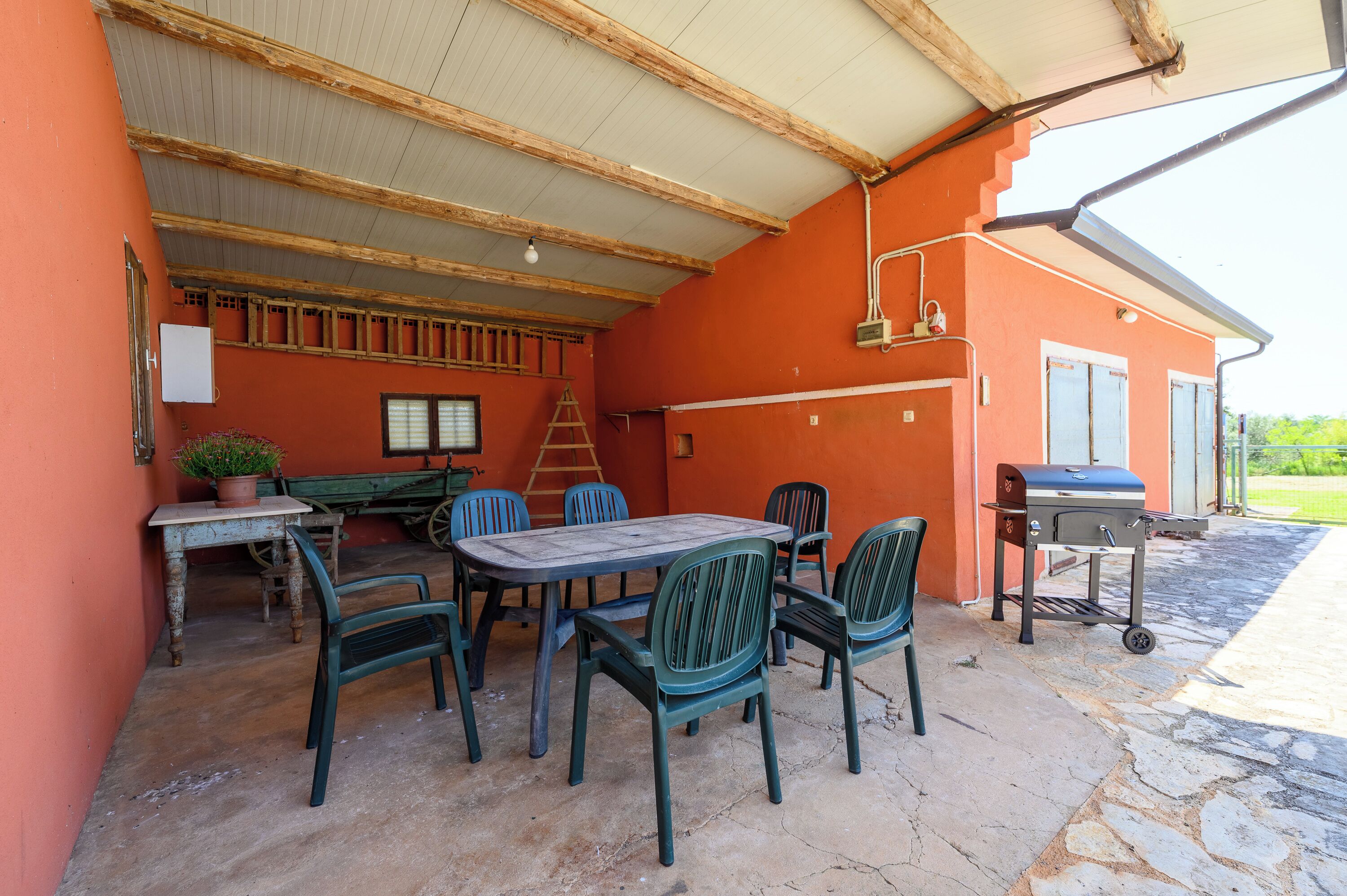 Um pátio externo com mesa e cadeiras, churrasqueira e uma escada encostada em um edifício com paredes alaranjadas.