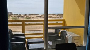 En balkon med fletstole og et bord, med udsigt over en strand med klitter og havet.
