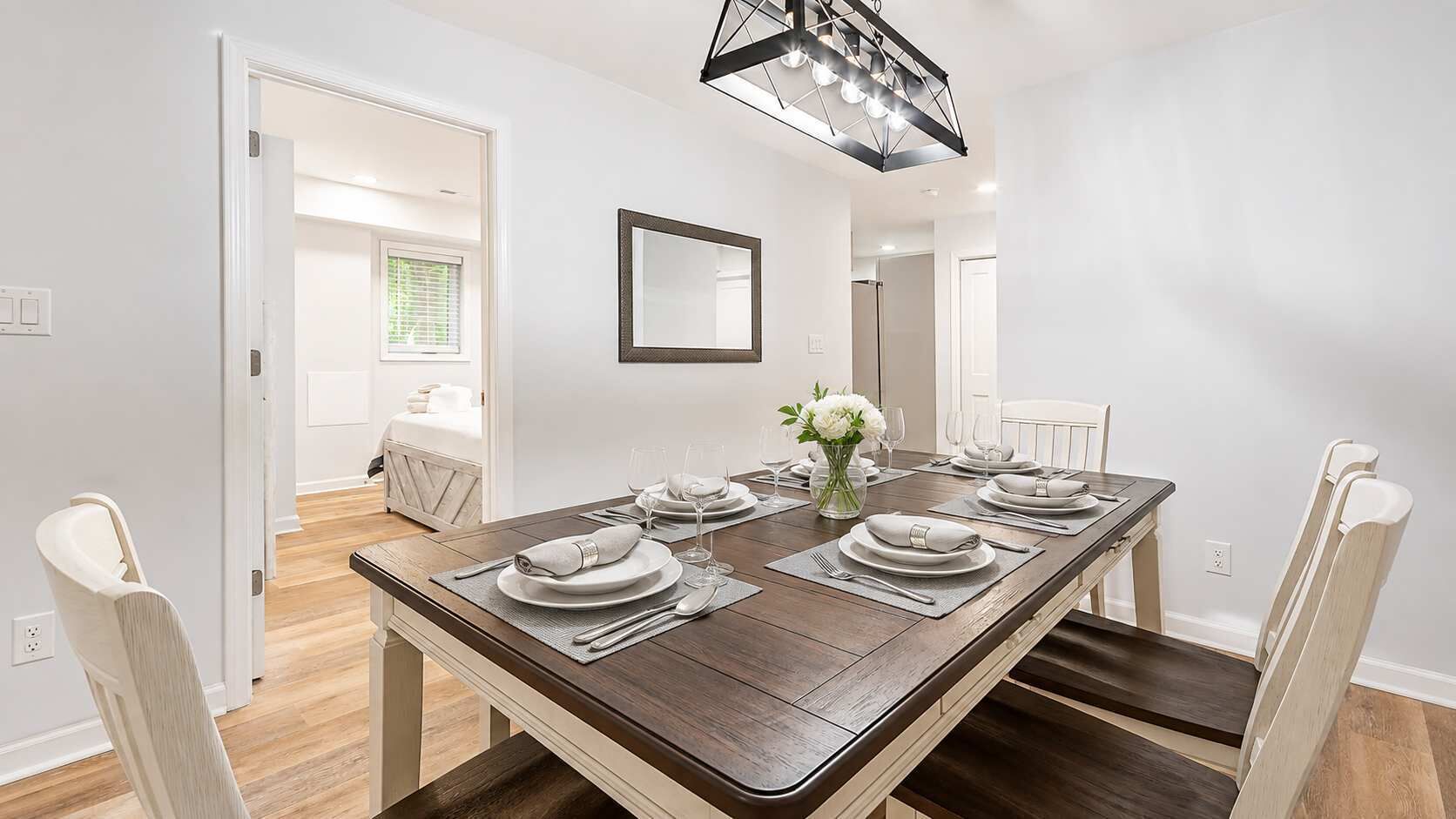 A dining room with a rectangular table set for four, white chairs, a vase with flowers, and a modern pendant light.