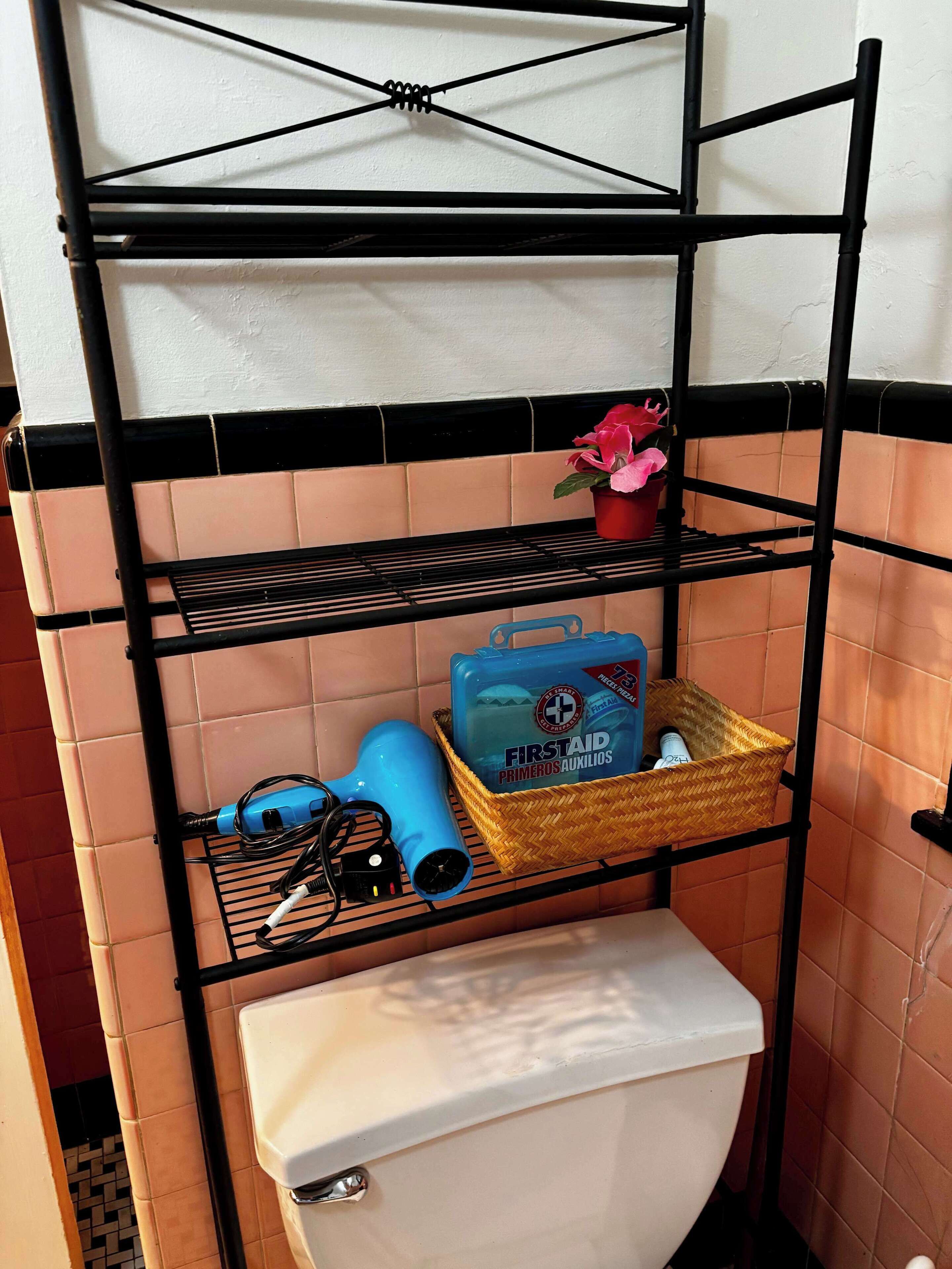 A bathroom with a black metal shelf unit above a toilet, holding a blue hairdryer, a basket, and a small potted plant.