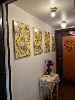 Un pasillo con arte floral en las paredes, una pequeña mesa con un florero y una puerta con un cartel de bienvenida.