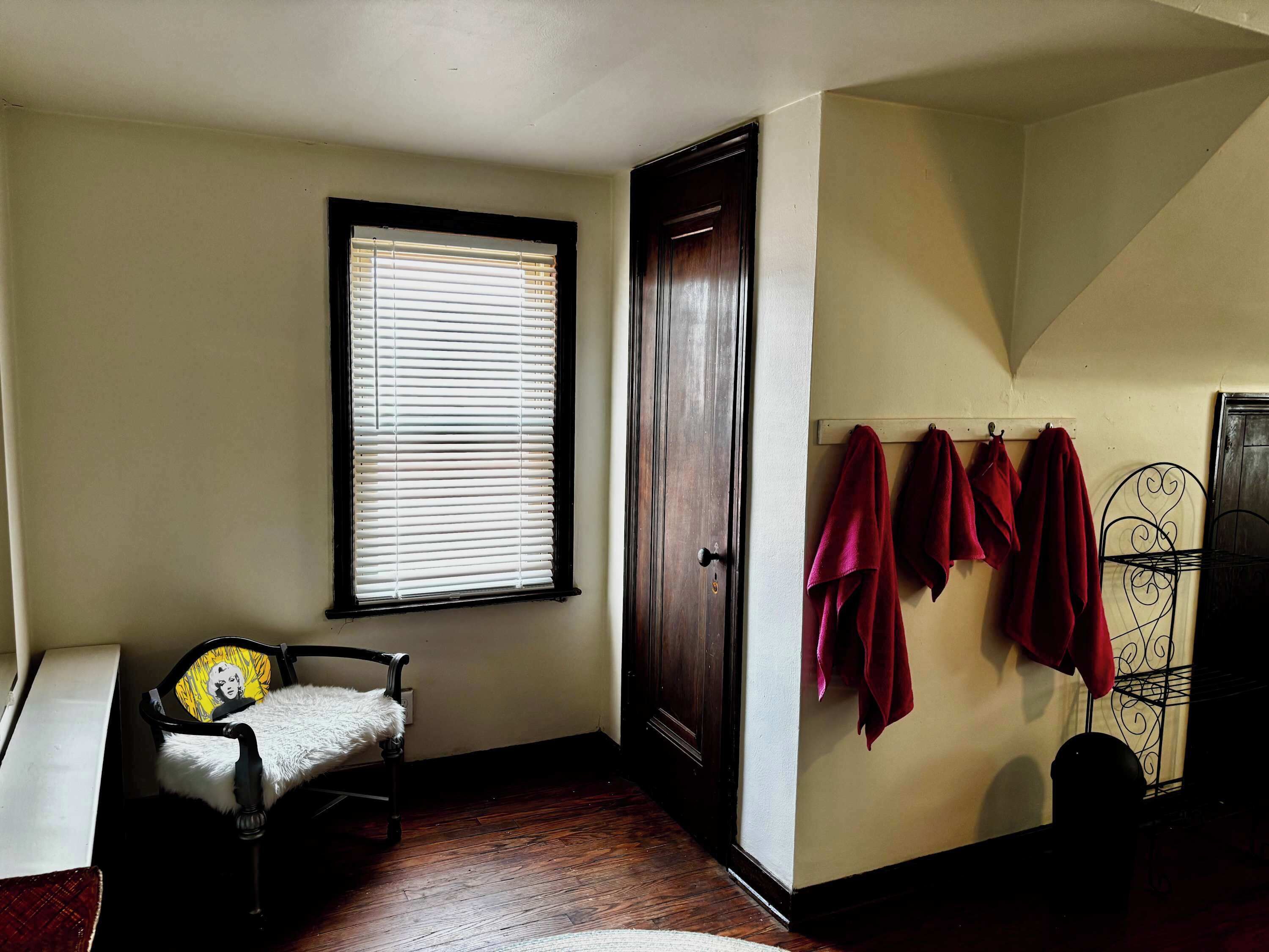 Ein Zimmer mit Holzboden, einem Sessel mit weißem Fellbezug, einem Fenster mit Jalousien, einer Tür und einer Wand mit roten Handtüchern.