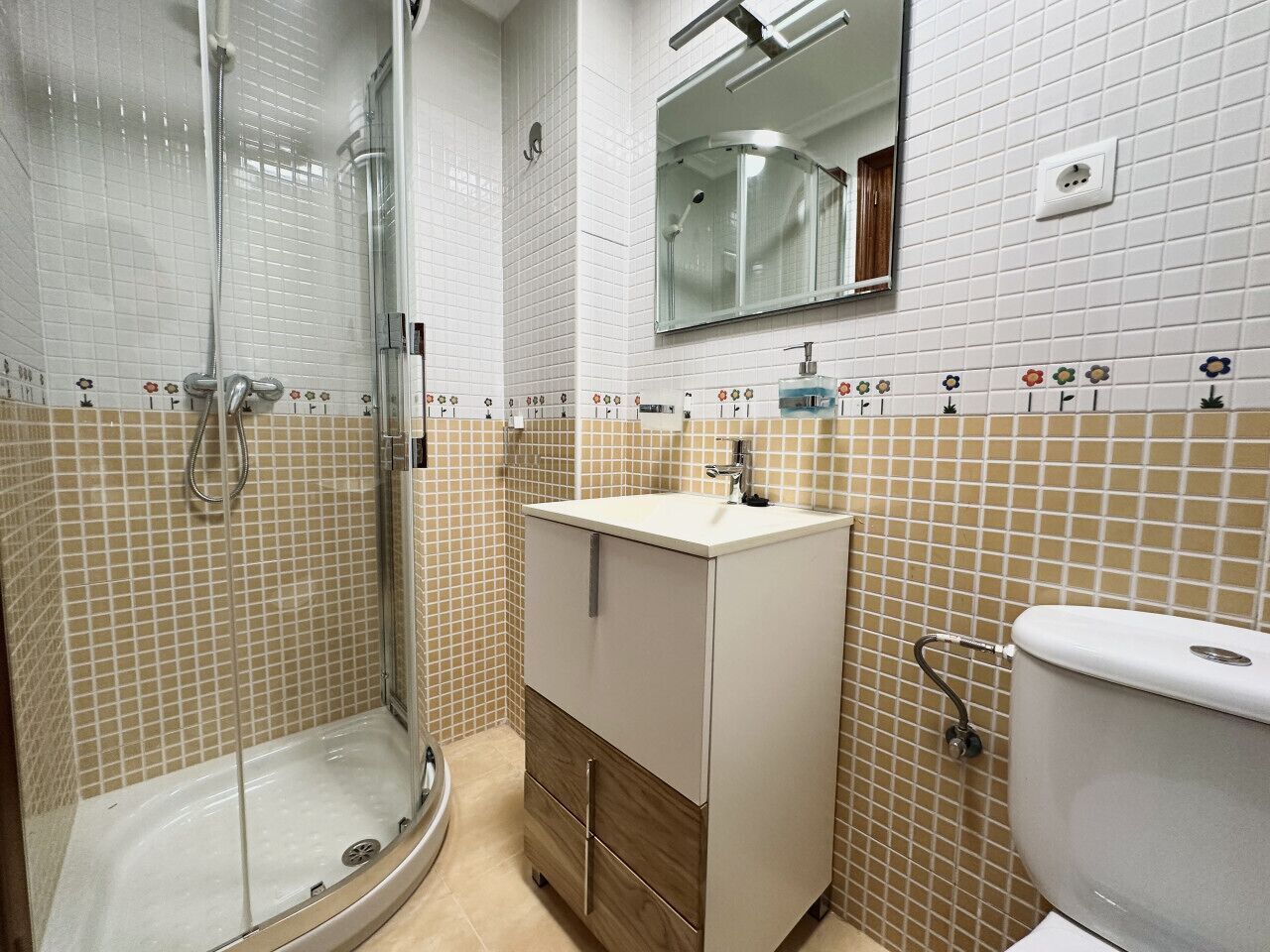 Et badeværelse med brusekabine i glas, vask med spejl og toilet.