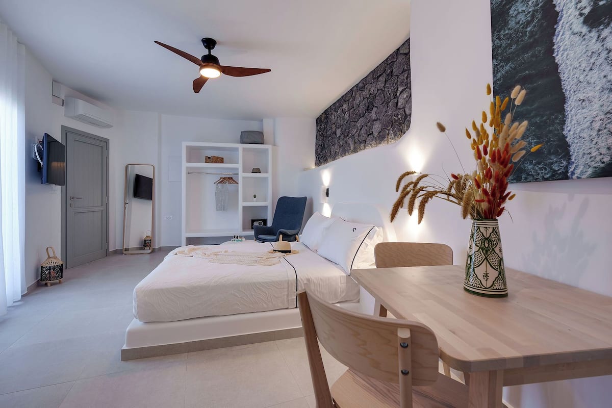 A modern hotel room with a bed, a desk, a chair, a ceiling fan, a TV, a wardrobe, and a wall-mounted shelf.