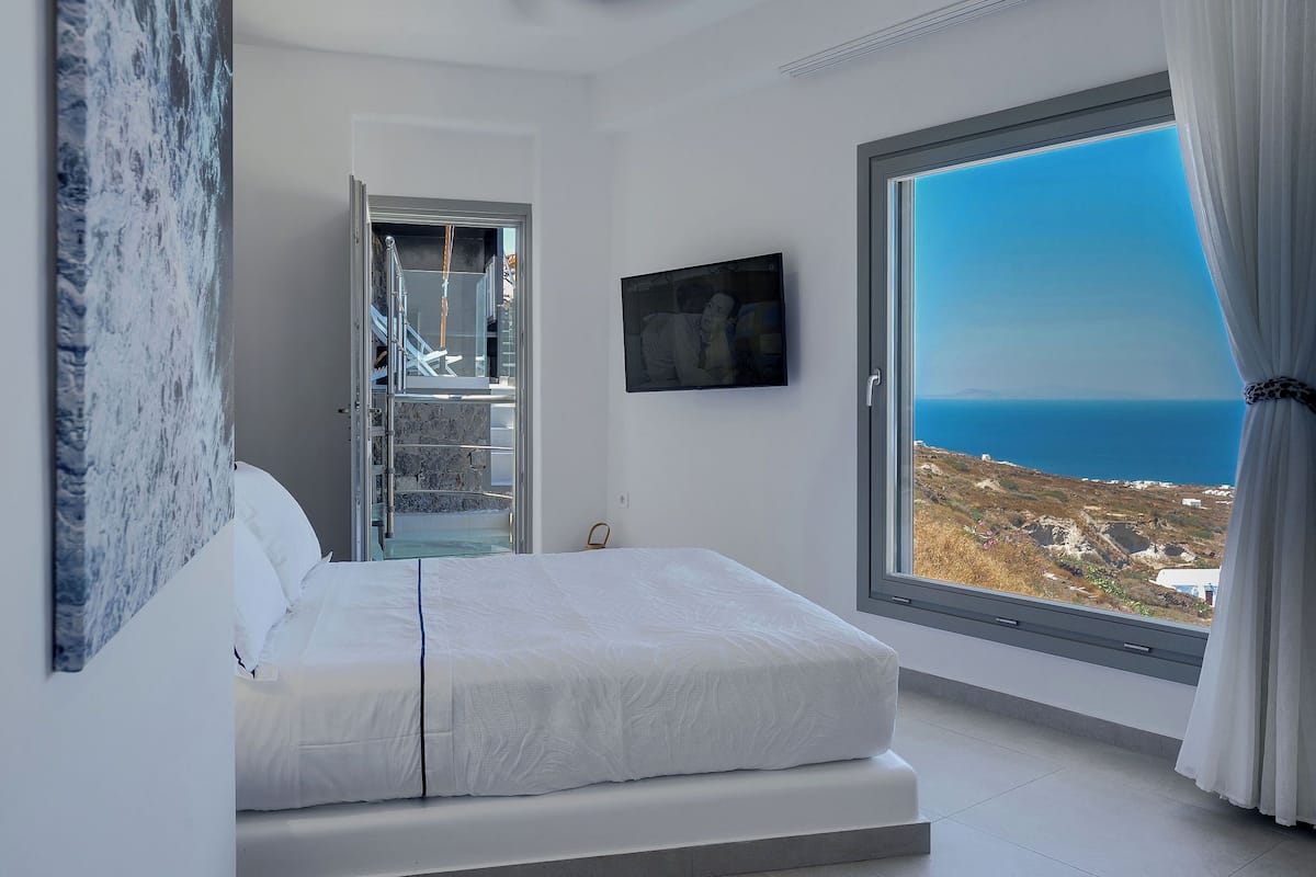 A bedroom with a bed, a large window with a view of the sea, and a painting on the wall.